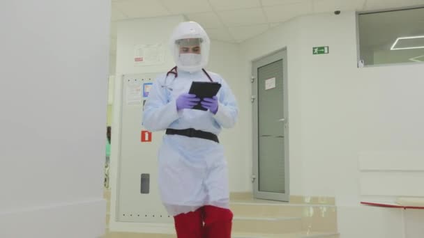 Doktor modern kliniğin koridoru boyunca yürür. Koruyucu kıyafetli bir doktor. Elinde tabletle bir doktor hastane koridorunda yürüyor.. — Stok video