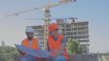 İnşaat alanındaki iki mühendis çizime bakıyor. Mühendisler çok katlı bir binanın önünde. Evin arka planında iki inşaatçı var.