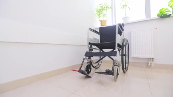 밝은 창문을 배경으로 빈 휠체어가 있다. 하얀 복도에 장애 인들을 위한 휠체어. 병원 복도에 장애가 있는 사람들을 위한 텅 빈 휠체어. — 비디오