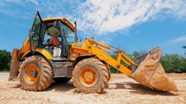 Büyük sarı bir kazıcı bir inşaat alanında araba sürüyor. Kazı tekerleri kapanıyor.