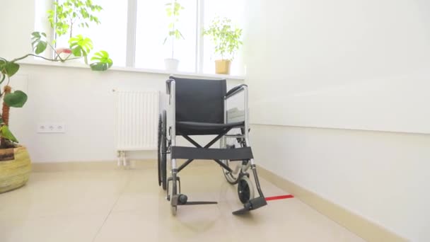 Empty wheelchair for people with disabilities in the hospital corridor. A wheelchair for people with disabilities in the white hallway. Empty wheelchair against the background of a bright window — Stock Video