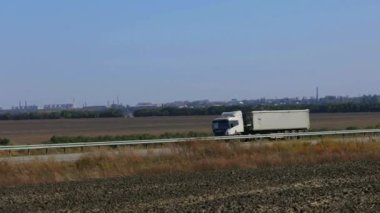 Güneşli bir havada beyaz bir araba otoban boyunca sürer. Otoyolda kamyon var.