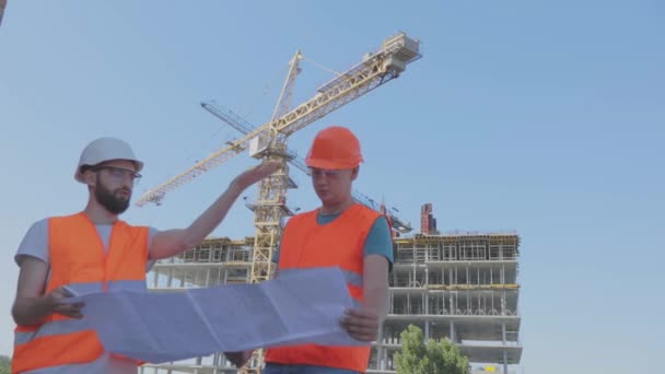 Inženýři před vícepodlažní budovou. Dva inženýři na staveništi se dívají na výkres. Dva stavitelé na pozadí domu — Stock video