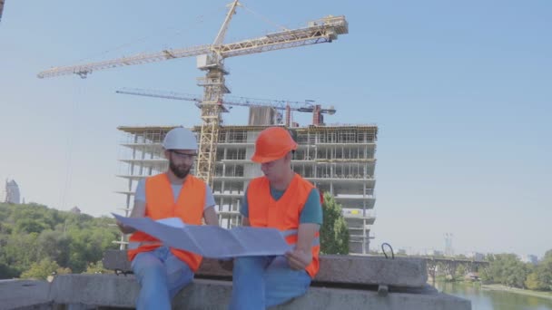 Dwóch inżynierów na placu budowy patrzy na rysunek. Inżynierowie przed wielopoziomowym budynkiem. Dwóch budowniczych na tle domu — Wideo stockowe