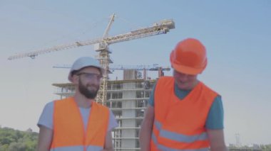 Mühendisler çok katlı bir binanın önünde. İnşaat alanındaki iki mühendis çizime bakıyor. Evin arka planında iki inşaatçı var.