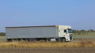 Güneşli bir havada beyaz bir araba otoban boyunca sürer. Otoyolda kamyon var.