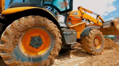 Büyük sarı bir kazıcı bir inşaat alanında araba sürüyor. Kazı tekerleri kapanıyor.