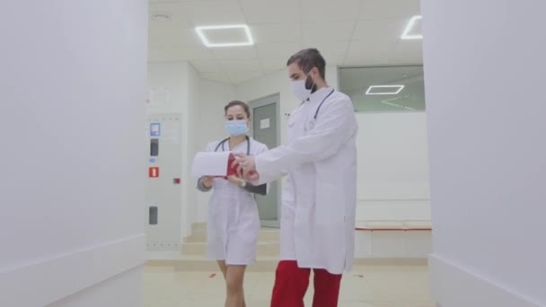 Bir doktor ve bir hemşire hastane koridorunda yürüyorlar. Bir doktor ve bir hemşire koridorda yürür ve konuşurlar. Hastane içi. — Stok video