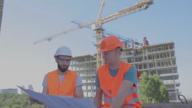 İnşaat alanındaki iki mühendis çizime bakıyor. Mühendisler çok katlı bir binanın önünde. Evin arka planında iki inşaatçı var.