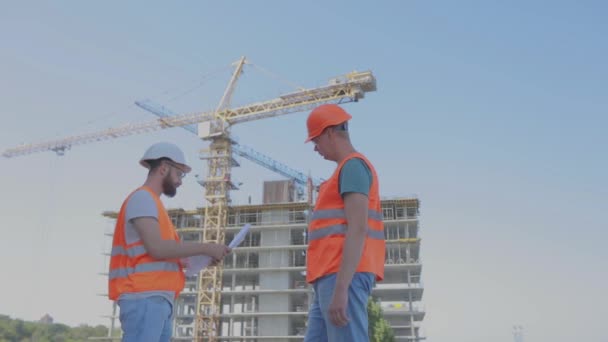 Dos ingenieros en un sitio de construcción están mirando el dibujo. Ingenieros frente a un edificio de varios pisos. Dos constructores en el fondo de la casa — Vídeos de Stock