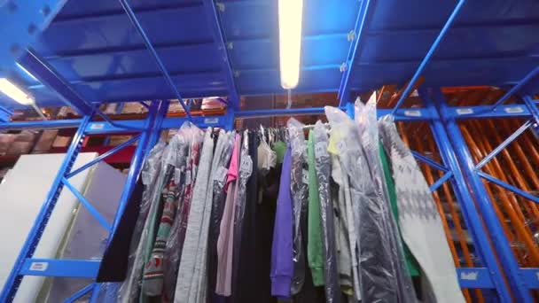 เสื้อผ้าถูกแขวนเป็นแถวในโกดัง คลังสินค้าพร้อมเสื้อผ้า เสื้อผ้าในโกดังของโรงงาน ภายในอุตสาหกรรม — วีดีโอสต็อก
