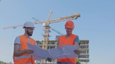 Mühendisler çok katlı bir binanın önünde. İnşaat alanındaki iki mühendis çizime bakıyor. Evin arka planında iki inşaatçı var.