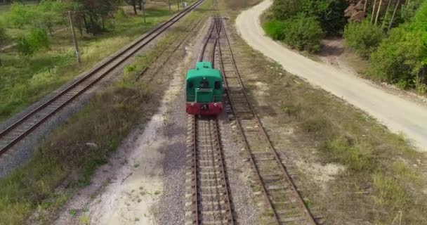 La piccola locomotiva si muove lungo la ferrovia nel bosco, la locomotiva verde si muove lungo la strada forestale vista aerea. Il movimento del treno viene filmato dal drone — Video Stock