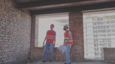 Evin arka planında iki inşaatçı var. Mühendisler çok katlı bir binanın önünde. İnşaat alanındaki iki mühendis çizime bakıyor..