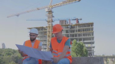 İki mühendis inşaat alanında bir çizim okuyorlar. Mühendisler, bir apartmanın arka planındaki çizimlere bakıyorlar.