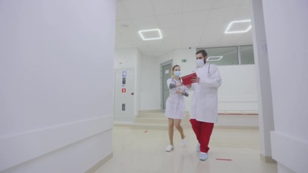 Doktorlar hastanenin parlak modern koridorları boyunca yürürler. Antiviral korumadaki doktorlar parlak hastane koridorlarında yürüyorlar. — Stok video