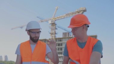 İki mühendis inşaat alanında bir çizim okuyorlar. Mühendisler, bir apartmanın arka planındaki çizimlere bakıyorlar.