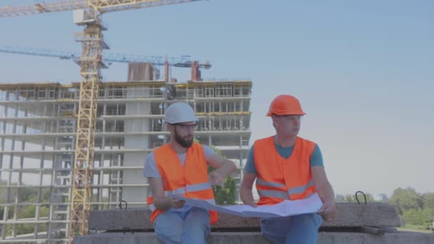 Инженеры перед многоэтажным зданием. Два инженера на строительной площадке смотрят на чертеж. Два строителя на заднем плане дома — стоковое видео