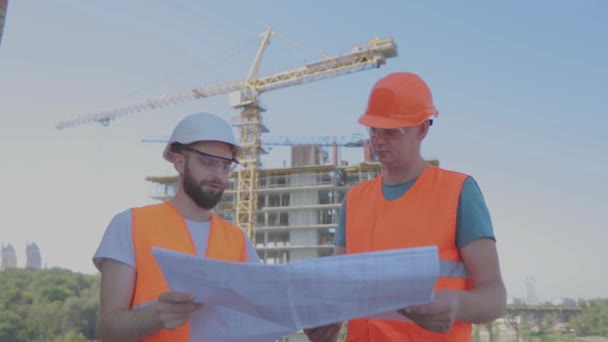 Ingenieurs voor een gebouw met meerdere verdiepingen. Twee ingenieurs op een bouwplaats kijken naar de tekening. Twee bouwers op de achtergrond van het huis — Stockvideo