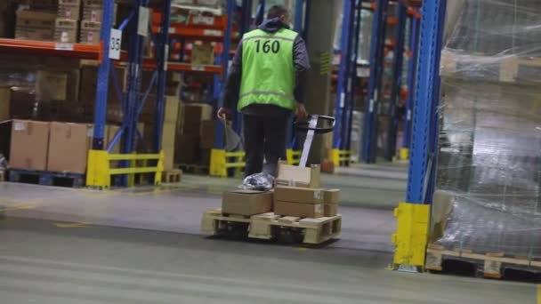 Muž ve velkém skladišti veze hydraulický vozík. Muž ve skladišti nese roklu. Dělník ve skladišti. Velký moderní sklad — Stock video
