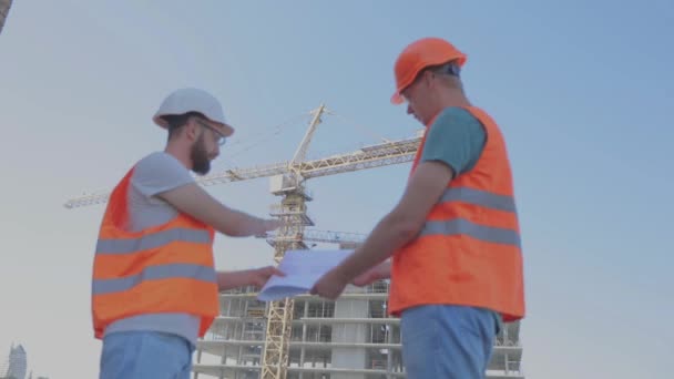 Engenheiros em frente a um edifício de vários andares. Dois engenheiros em um canteiro de obras estão olhando para o desenho. Dois construtores no fundo da casa — Vídeo de Stock