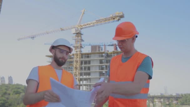 Dwóch inżynierów na placu budowy patrzy na rysunek. Inżynierowie przed wielopoziomowym budynkiem. Dwóch budowniczych na tle domu — Wideo stockowe