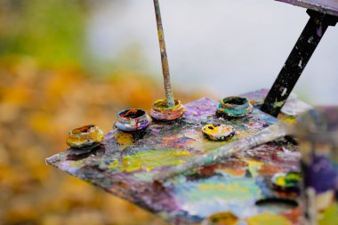 Güneşli bir sonbahar gününde bir manzara çizerken bir ressam ve renk paleti ile sığ alan derinliği (seçici odak) resmi.