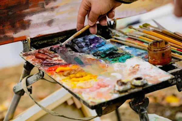 Güneşli bir sonbahar gününde bir manzara çizerken bir ressam ve renk paleti ile sığ alan derinliği (seçici odak) resmi.