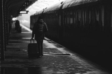 Siyah-beyaz ve (kara film, atmosferik) görüntüde, kar yağışlı bir kış gününde tren istasyonundan ayrılmaya hazır bir bavul taşıyan bir adam görülüyor..