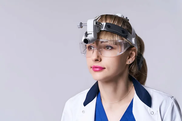 Sebészeti fényszóró fényszórót és védőszemüveget viselő, magabiztos ENT orvos. Szoros portré női otolaryngologist vagy fej-nyak sebész világosszürke háttér. — Stock Fotó