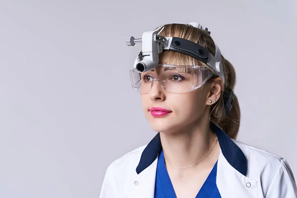 Médico otorrinolaringologista confiante vestindo luz da cabeça do farol cirúrgico e óculos de proteção. Retrato de close-up de otorrinolaringologista feminino ou cirurgião de cabeça e pescoço sobre fundo cinza claro. — Fotografia de Stock