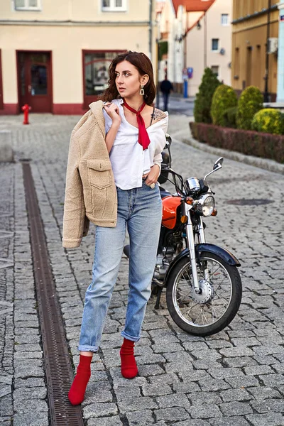 Jovem mulher bonita em vermelho tecido de malha calcanhar calcanhar calça calça jeans azul e casaco de pelúcia posando perto de uma motocicleta em uma rua — Fotografia de Stock