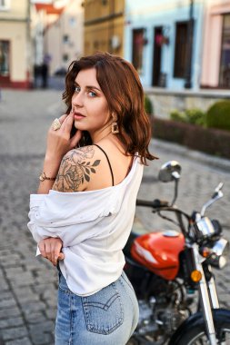 Kot pantolonlu ve beyaz gömlekli genç bir kadın sokakta motosikletin yanında poz veriyor.