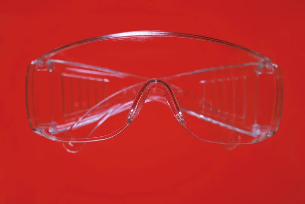 Protective glasses on vivid red background, healthcare and safety concept. Plastic safety glasses. Personal protective equipment. Plastic goggles, transparent glasses safety at work