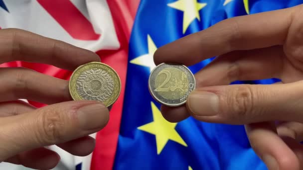 英国と欧州連合の旗を背景に 2ポンド硬貨と2ユーロ硬貨が手に入ります イギリスはブレグジット後 金利だ 貨幣交換の概念 4Kビデオ — ストック動画