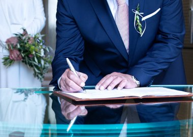 Koyu mavi takım elbiseli yaşlı damat masada evlilik sözleşmesi imzalıyor. Adamın eli kalem tutuyor ve kağıt imzalıyor.