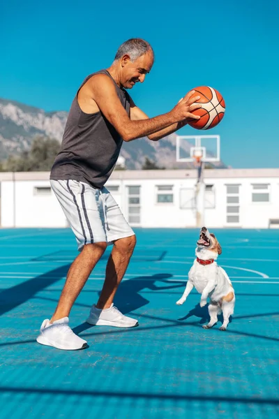 Portrait of elderly turkish cypriot man playing basketball with a small cute active jumping dog jack russel terrier on playground outdoor
