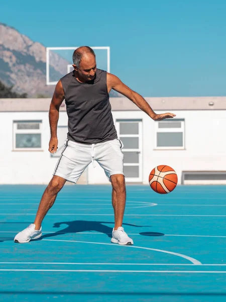 Adult senior man playing a sport ball on blue basketball court at summer sunny day against backdrop of mountains and sky