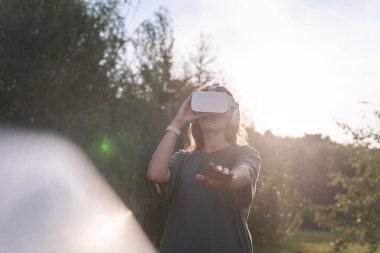 Elleriyle sanal gerçeklik gözlüğü takan bir kız. Bir elini kameraya uzatıyor, güneş ışığının yanına..