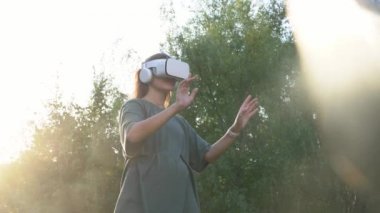 Sanal gerçeklik gözlüklü bir kız yeşil ağaçların arkasında duruyor. Ellerini havada hareket ettiriyor, güneş ışığını yakalamaya çalışıyor. Gelecek kavramı..