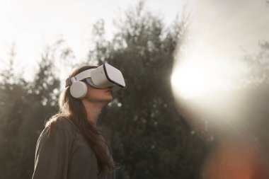 Sanal gerçeklik gözlüğü takan kız gözlüğü elleriyle tutuyor. Yeşil ağaçların arka planında. Yakınlarda bir güneş ışığı var..