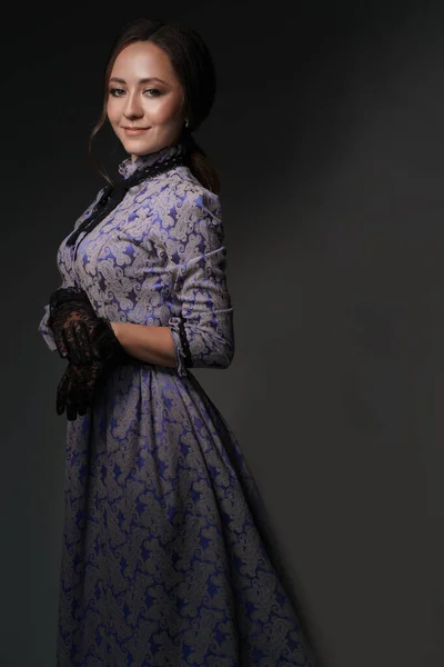 Una niña de pie en un vestido azul en el estilo de la década de 1900, en un vestido retro, un sombrero sobre un fondo gris. Moda histórica del siglo XIX Fotos de stock libres de derechos