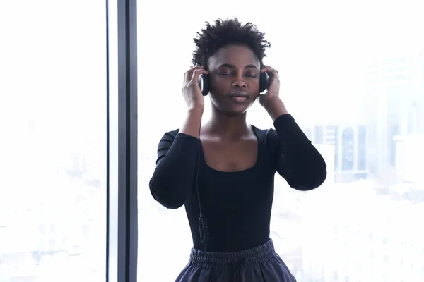 Uma mulher afro-americana ouve música com auscultadores. Uma jovem mulher de uniforme esportivo no fundo de uma janela — Fotografia de Stock