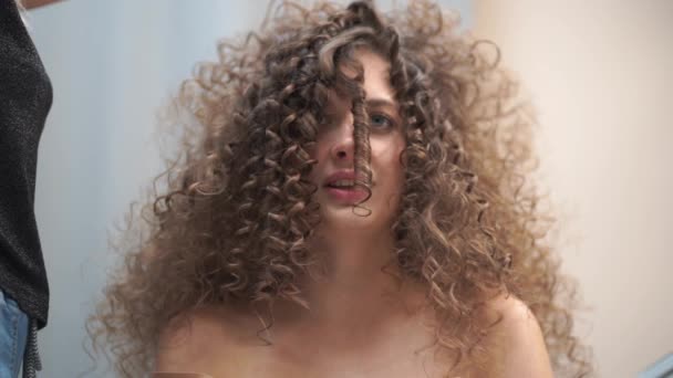 Movimento lento. Close-up de uma mulher bonita com cabelo encaracolado, seu cabelo está sendo feito, ela sopra em um fio de cabelo, olhando para a câmera — Vídeo de Stock