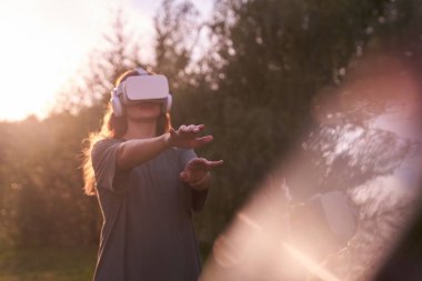 Elleriyle sanal gerçeklik kaskı tutan bir kız. Bir elini kameraya uzatıyor, güneş ışığının yanında..