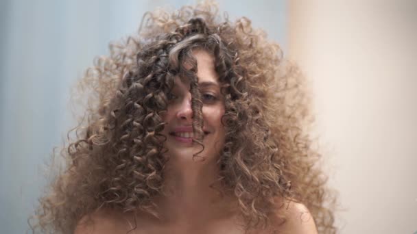 Langsom bevægelse. Close-up af en smuk kvinde med krøllet hår, hendes hår bliver gjort, hun blæser på en streng af hår, ser ind i kameraet – Stock-video