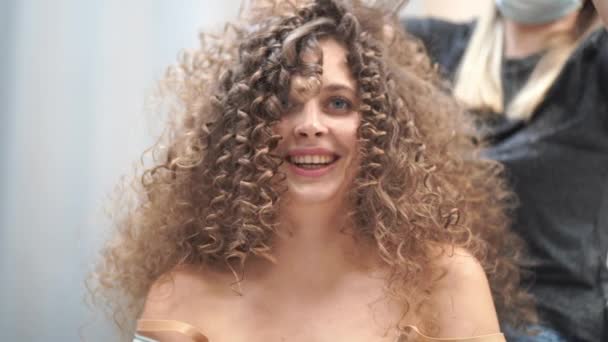 Närbild av en vacker kvinna med lockigt hår, hennes hår håller på att göras, hon blåser på en hårstrå tittar på kameran — Stockvideo
