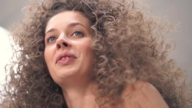 Aşağıdan, kıvırcık saçlı, güzel bir genç kadının yakın plan çekimi, başını çeviriyor ve konuşuyor. Efendi ona makyaj yapıyor.