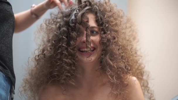 Au ralenti. Gros plan d'une belle jeune femme aux cheveux bouclés, elle regarde la caméra sourire, le maître fait ses cheveux — Video