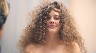 Kıvırcık saçlı güzel bir kadının yakın çekimi, saçları yapılırken, kameraya bakarak bir saç teline üflüyor.